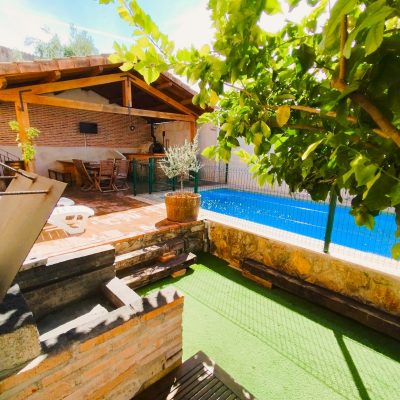 Patio de una casa rural rústica con paredes de ladrillo visto, una zona de porche de madera y una pequeña piscina rodeada de plantas.