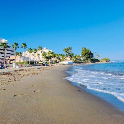 Playa de arena fina junto a un complejo de apartamentos turísticos y palmeras bajo un cielo azul despejado en Estepona.